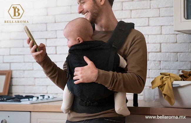 Hombre recargado en un mueble de cocina cargando a un bebé con ayuda de una mochila portabebés.