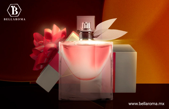 Perfume La Vie Est Belle de Lacome, frasco color rosa y con detalle de moño en la tapa, en frente de caja de regalo y sobre un fondo negro y naranja.