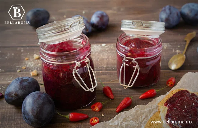 Dos frascos de vidrio con conservas artesanales de mermelada oscura rodeados de ciruelas frescas y chiles rojos sobre una mesa.