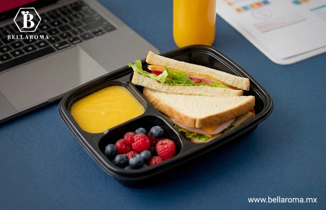 Contenedor de plástico con un desayuno para llevar que incluye sándwiches triangulares, frutos rojos y aderezo junto a una laptop.
