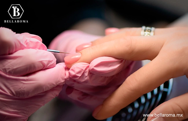 Manicurista utilizando guantes rosas y un pincel fino para aplicar esmalte de uñas en gel a una clienta.