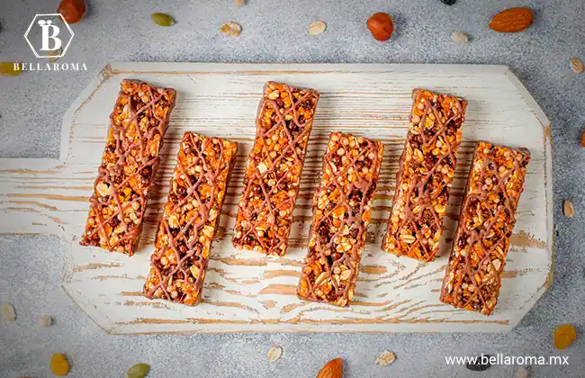 Barras de cereales y snacks saludables con hilos de chocolate sobre una tabla blanca de cocina rodeada de frutos secos.
