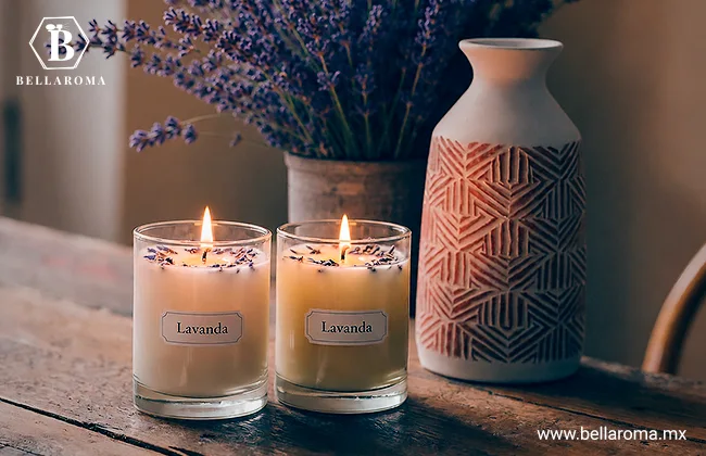 Dos velas artesanales encendidas en vasos de vidrio con etiquetas que dicen "Lavanda", colocadas sobre una mesa rústica de madera junto a un jarrón.